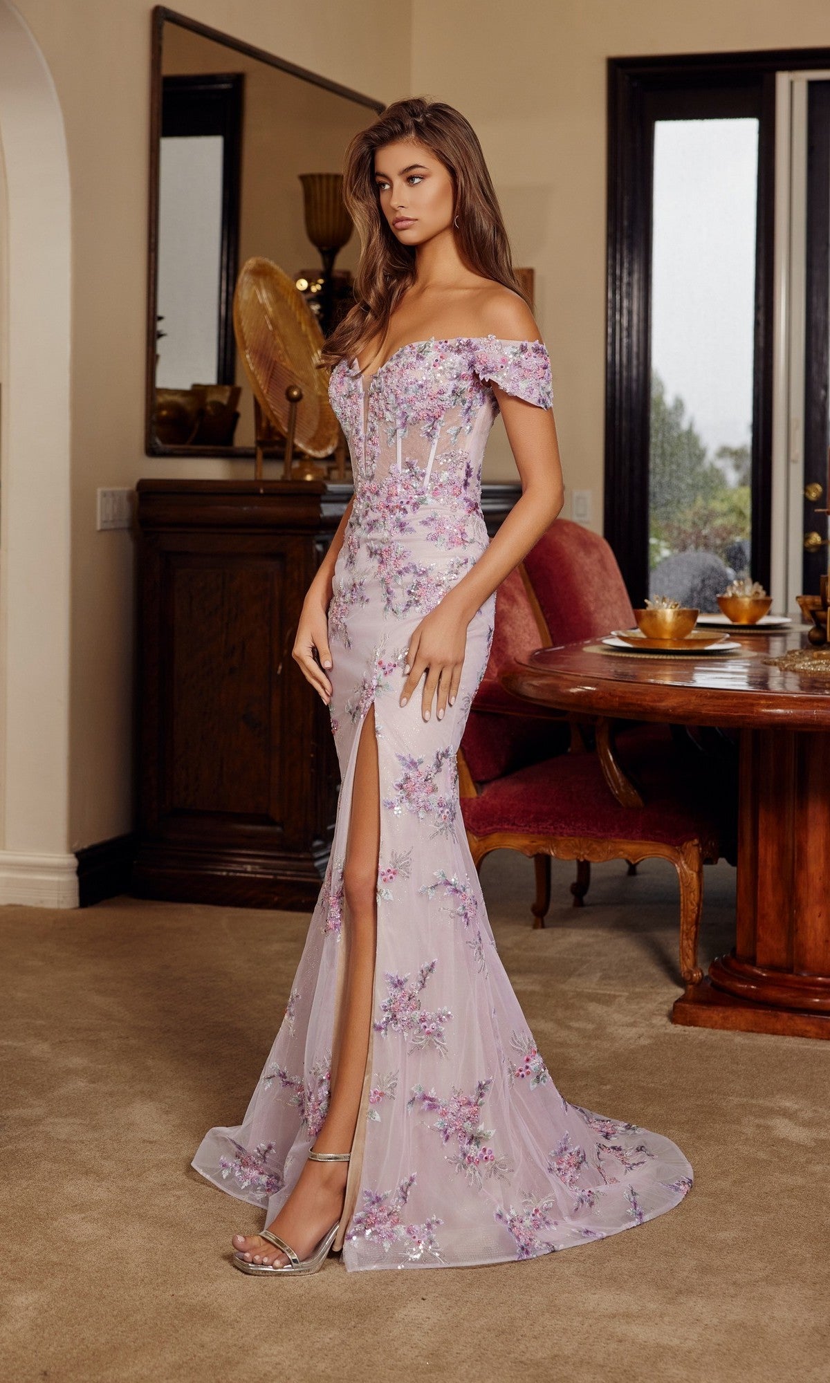 Woman in a Nox Anabel C1829 long prom dress featuring off-the-shoulder style, sequin floral embroidery, and a thigh-high slit, standing indoors on carpet near a wooden dining table and chairs.